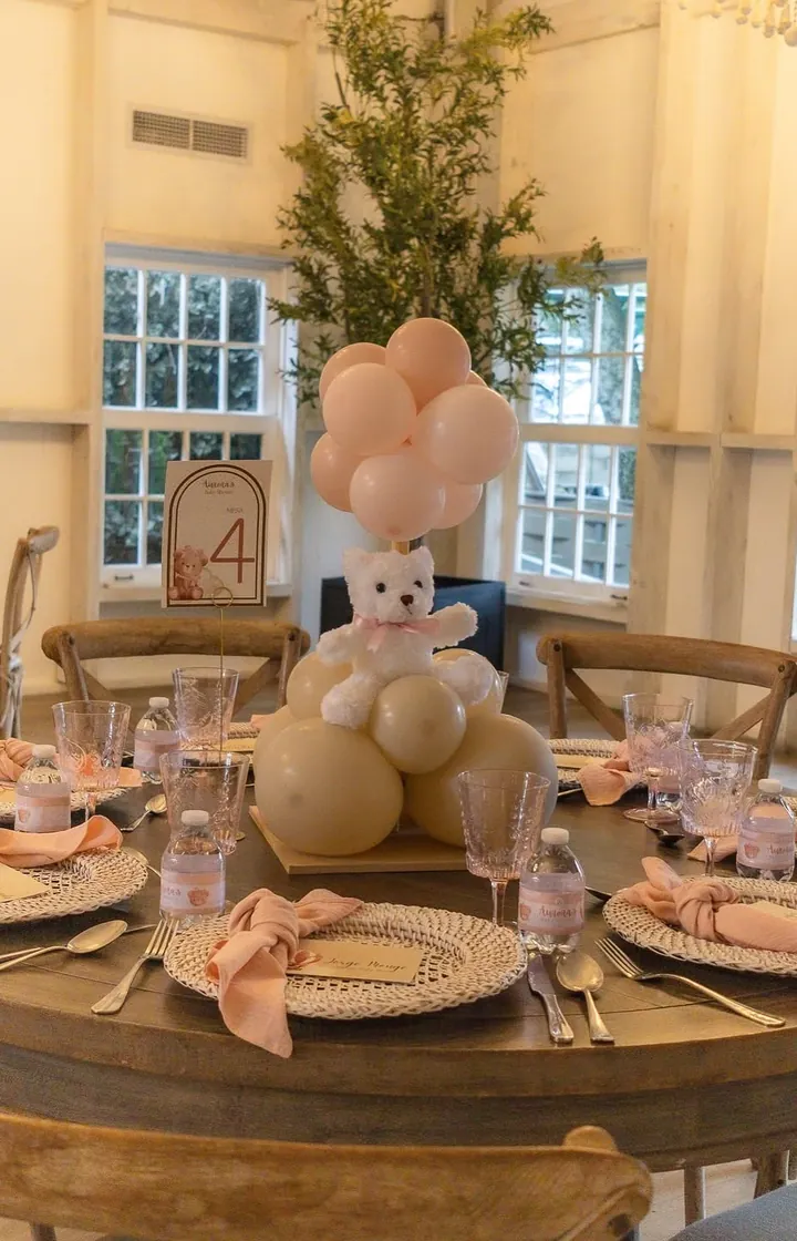 Mesa redonda de un baby shower con un oso de peluche, globos color arena y servilletas rosadas