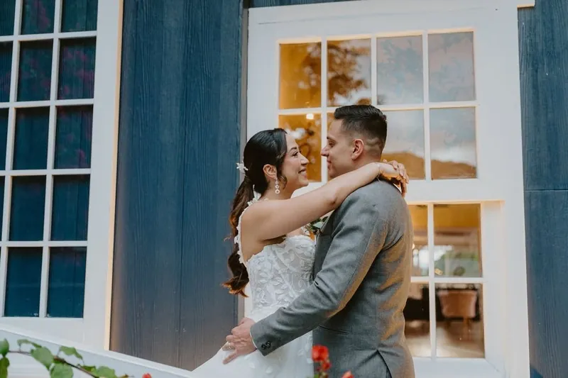 Novios abrazados frente a la pared azul de la casa y una ventana iluminada