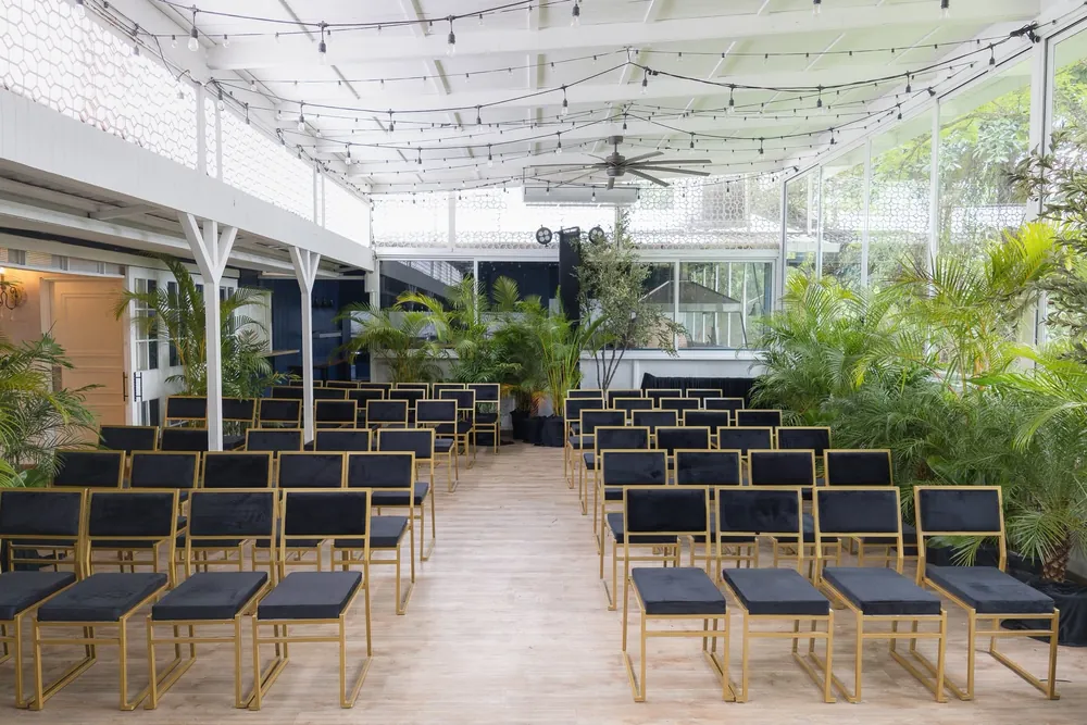 Espacio techado con ventanales hacia el jardín, plantas y filas de sillas negras